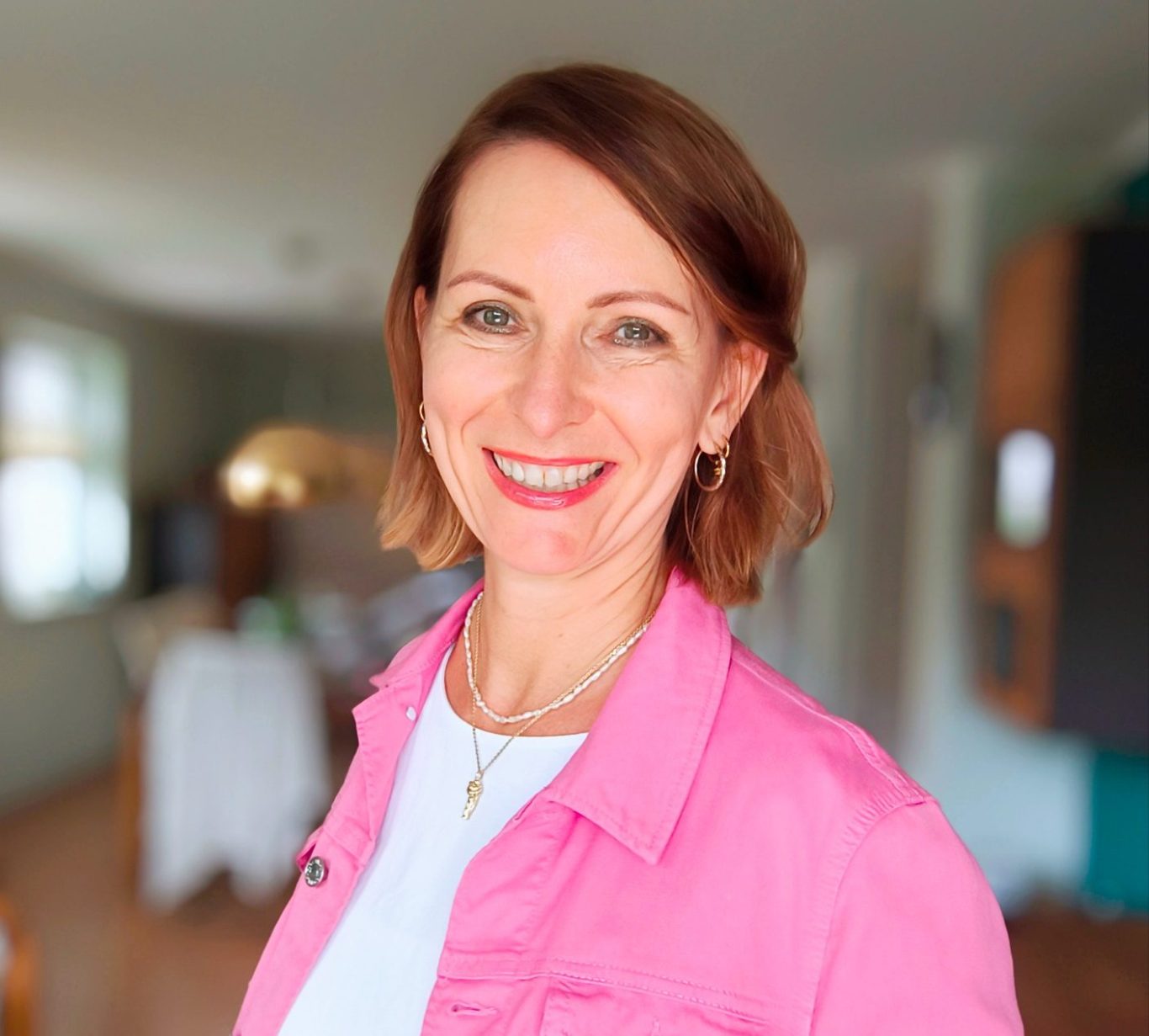 Frau mit kurzem, braunem Haar und rosa Jacke lächelt freundlich. HellerInnenraum im Hintergrund.