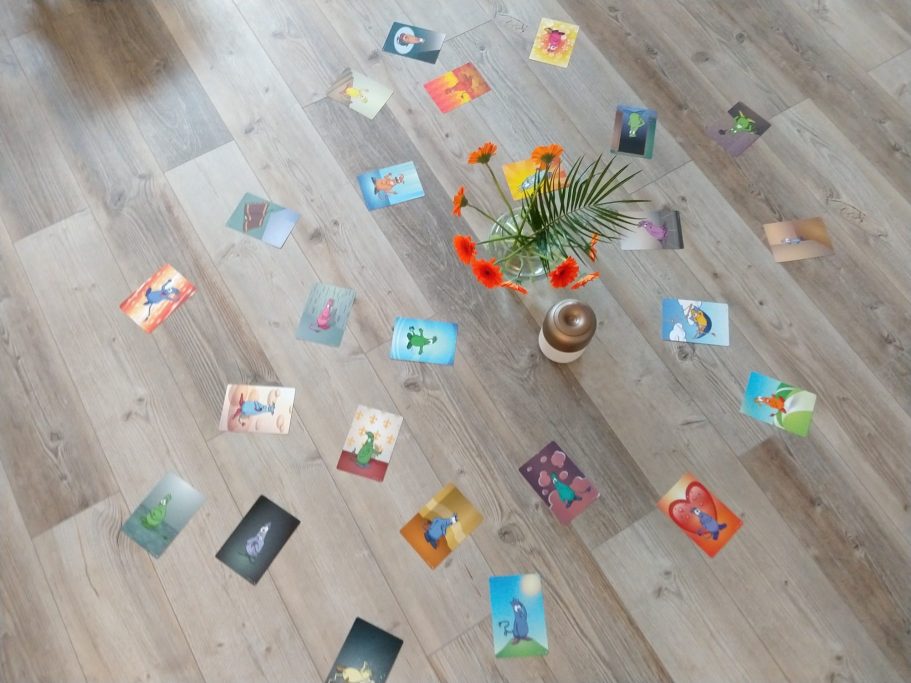 Bunte Karten liegen auf einem Holzboden um eine Vase mit orangefarbenen Blumen.